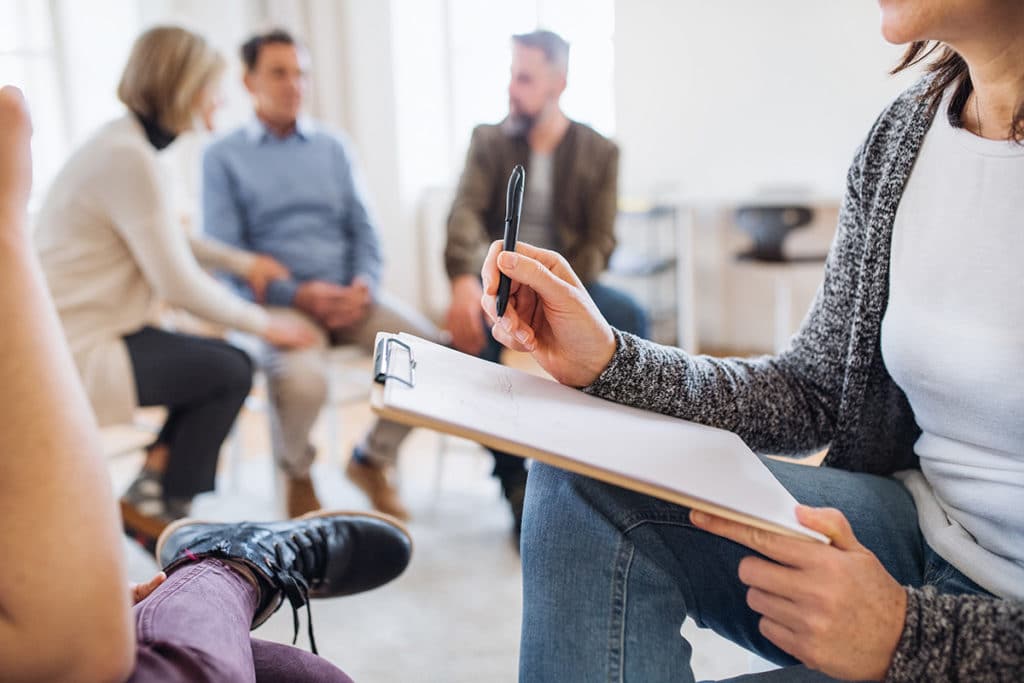 counselor in group therapy with clipboard talks to single patient about crystal meth vs meth while other patients talk in background