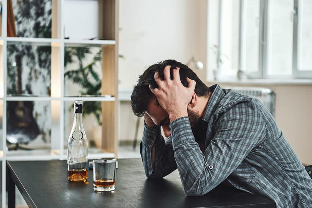 man runs his hands in his hair while looking at alcohol on the table and trying to avoid drinking on the job